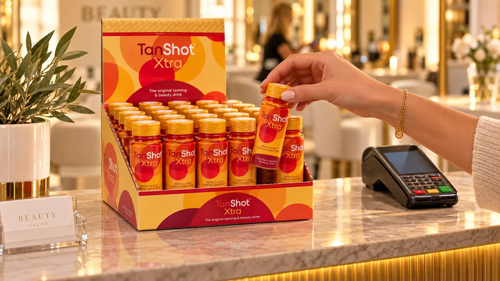 A case of TanShot Xtra open as a counter display on a salon reception desk, a client taking a bottle